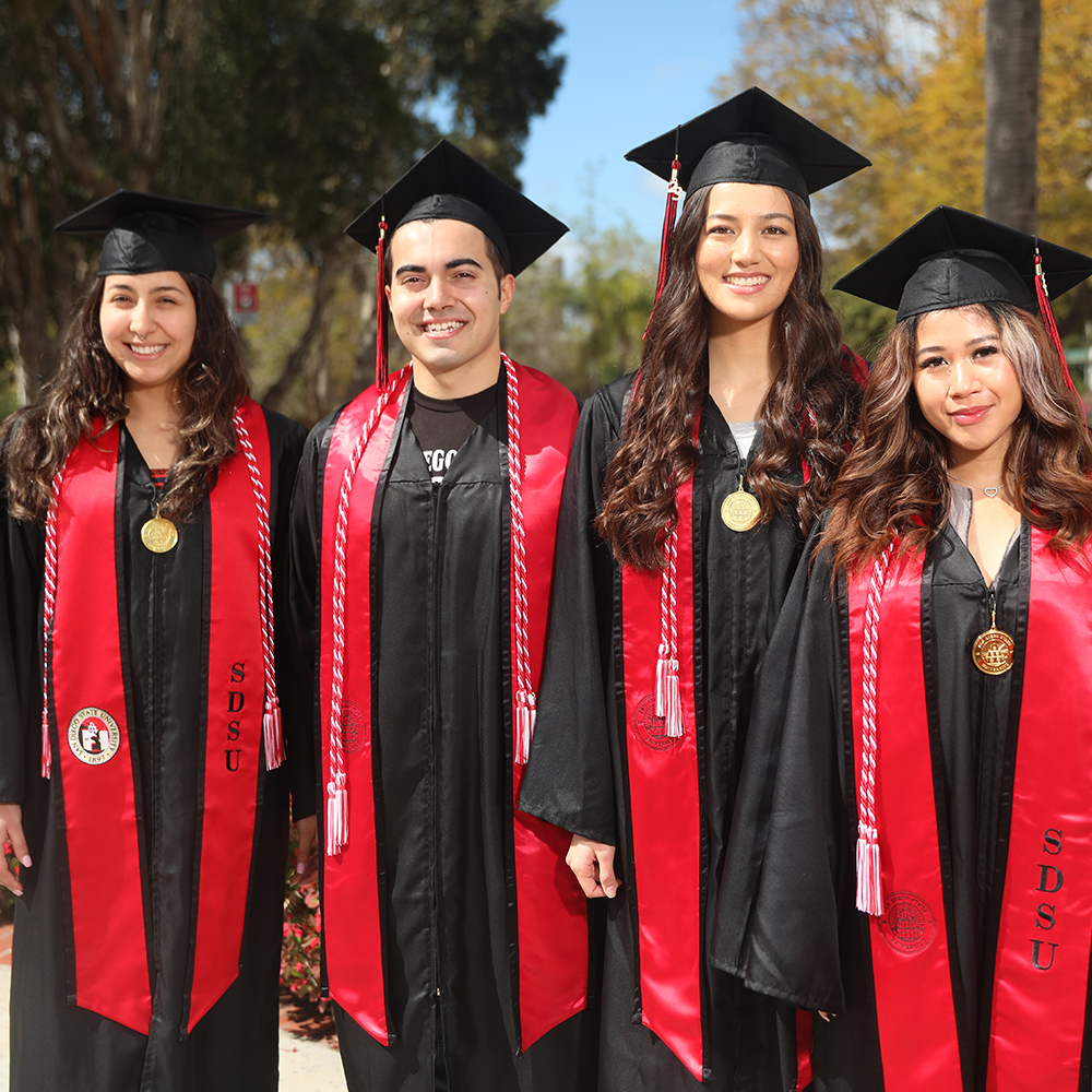Four SDSU students in their black graduation gown, red sashes, black caps, and red and black tassels. Posed outside in the sun.