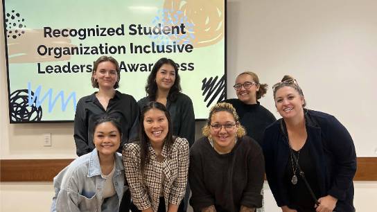  a group of women stand and crouch in front of a presentation screen that says Recognized Student Organization Inclusive Leadership Awareness
